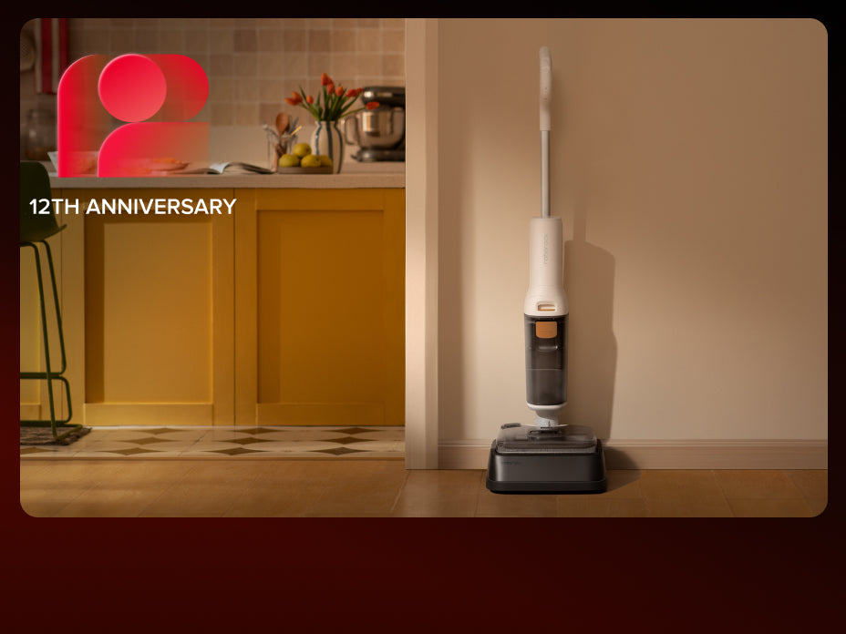 A person using a white Roborock F25 GT wet/dry vacuum to clean up a cracked egg and spill on a wooden kitchen floor, with a small kitten watching nearby