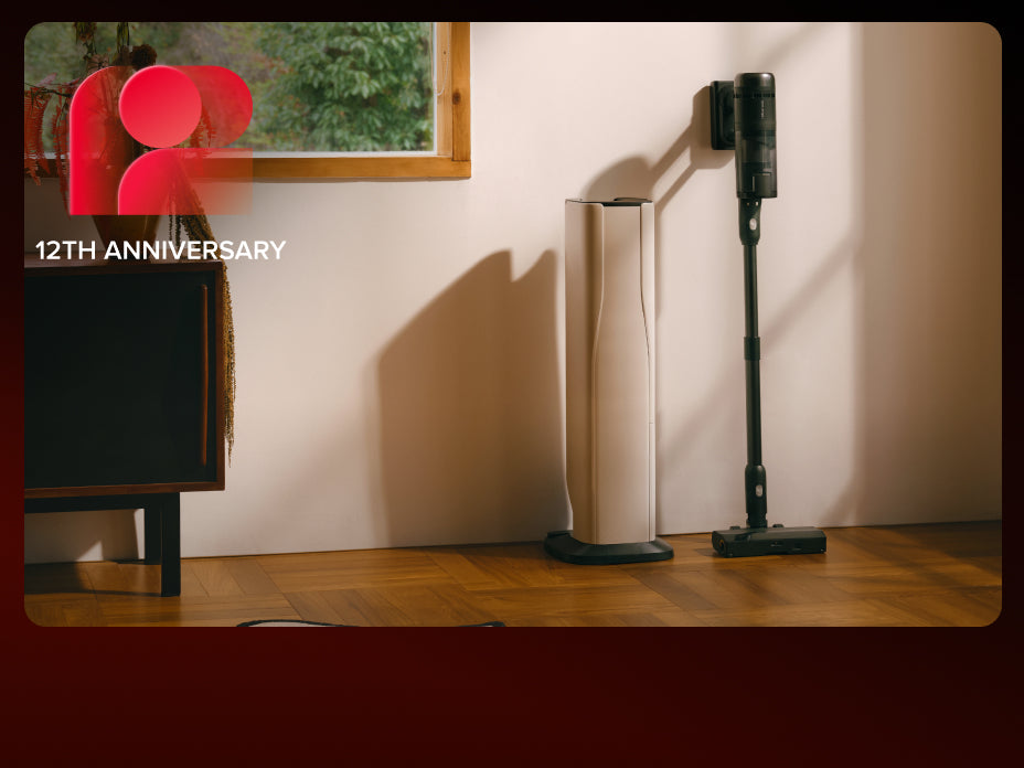 Black Roborock H60 Hub Ultra cordless stick vacuum leaning against a white wall next to its docking station on a wooden floor