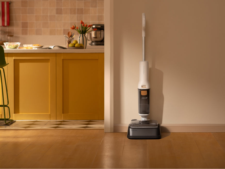 A person using a white Roborock F25 GT wet/dry vacuum to clean up a cracked egg and spill on a wooden kitchen floor, with a small kitten watching nearby